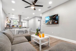 A modern living room with a gray sectional sofa, a white coffee table with fruit and drinks, a ceiling fan, and a kitchen in the background. - Remodeled 3BR, 6 Min to Busch Gardens | Fenced Yard, Monthly Stays Welcome (Tampa)