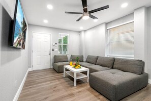 A modern living room with a sectional sofa, a small white table with fruit, a ceiling fan, and a TV mounted on the wall. - Remodeled 3BR, 6 Min to Busch Gardens | Fenced Yard, Monthly Stays Welcome (Tampa)