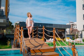 A person standing on a wooden deck by a pool with lounge chairs and a building in the background. - Elysium Deluxe Suites  (Antalya)