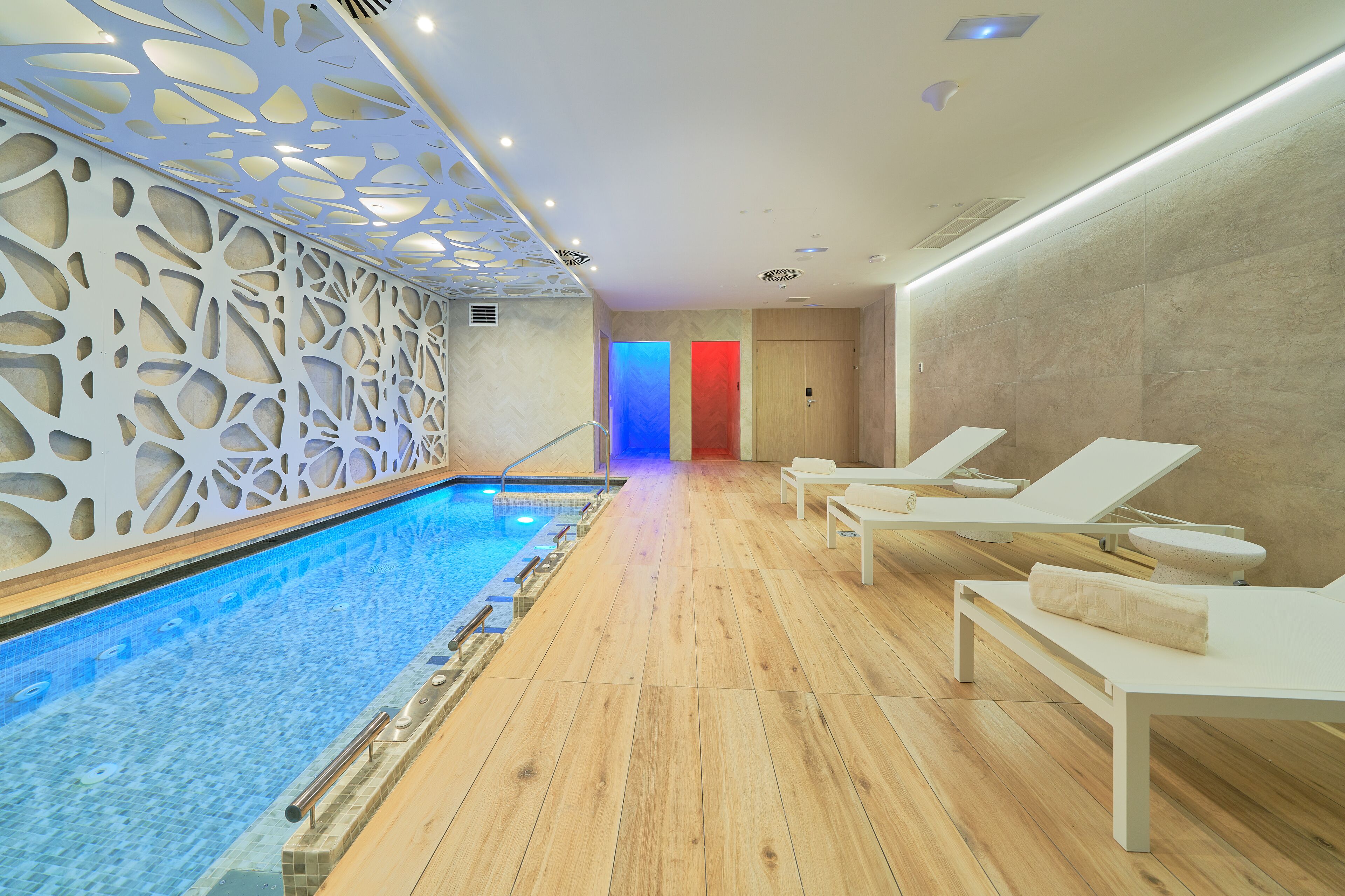 A modern indoor pool area with wooden flooring, lounge chairs, and a decorative ceiling.