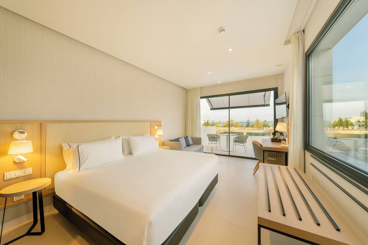 A modern hotel room with a large bed, a desk with a chair, a small table, and a large window offering a view of the city.