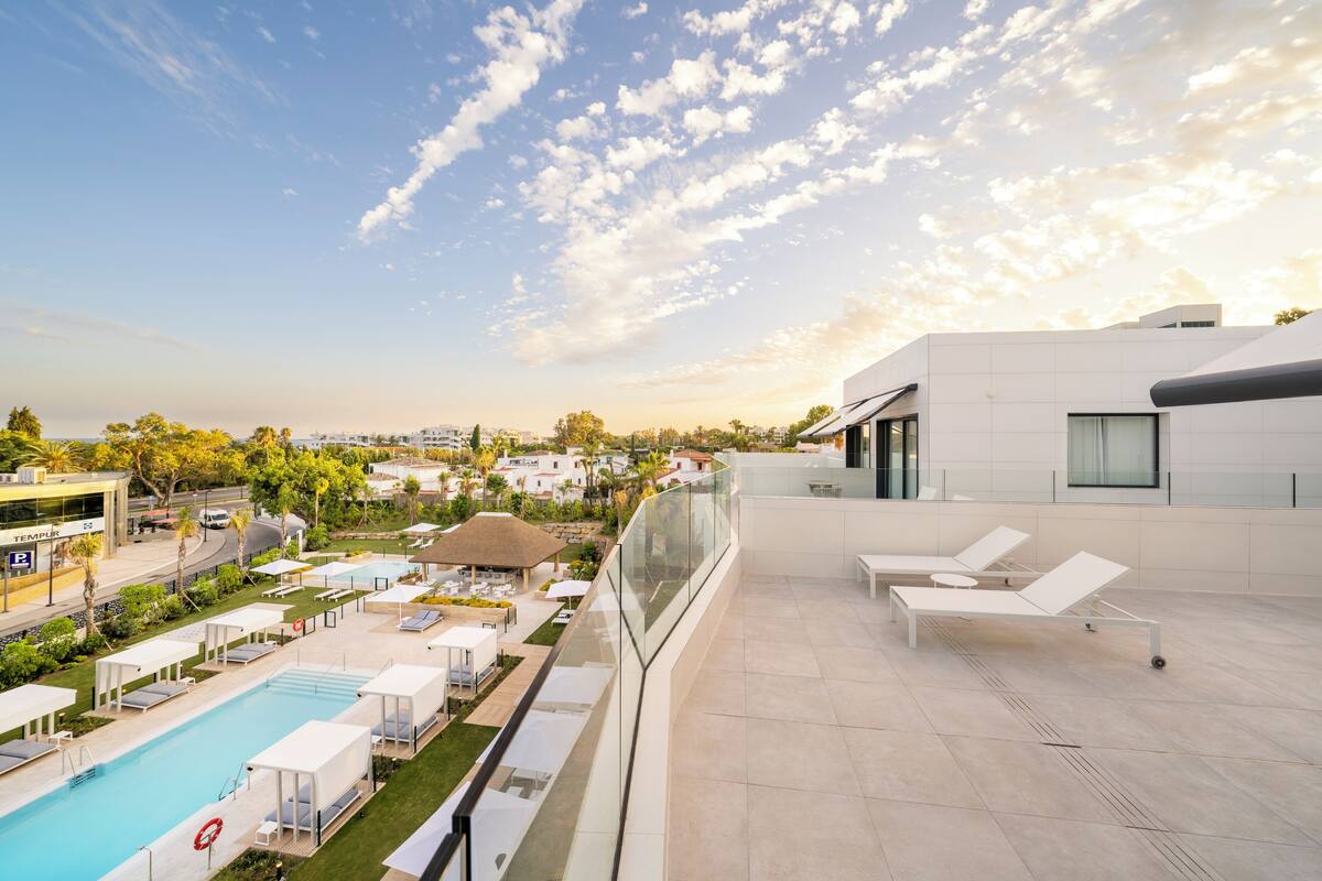 A rooftop pool area with lounge chairs, a swimming pool, and a glass balustrade overlooking a residential area.