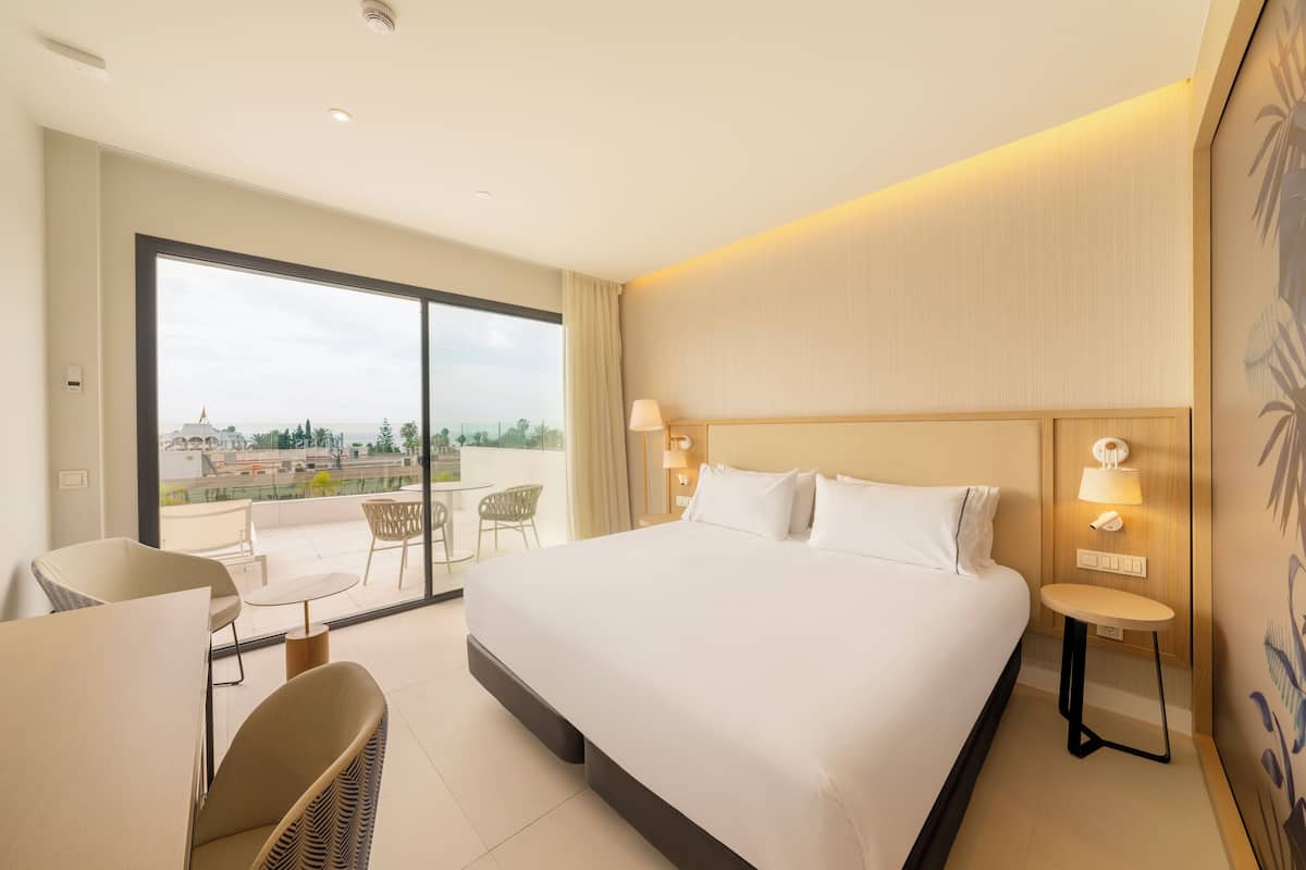 A hotel room with a large bed, a small table, a chair, and a balcony with a view.