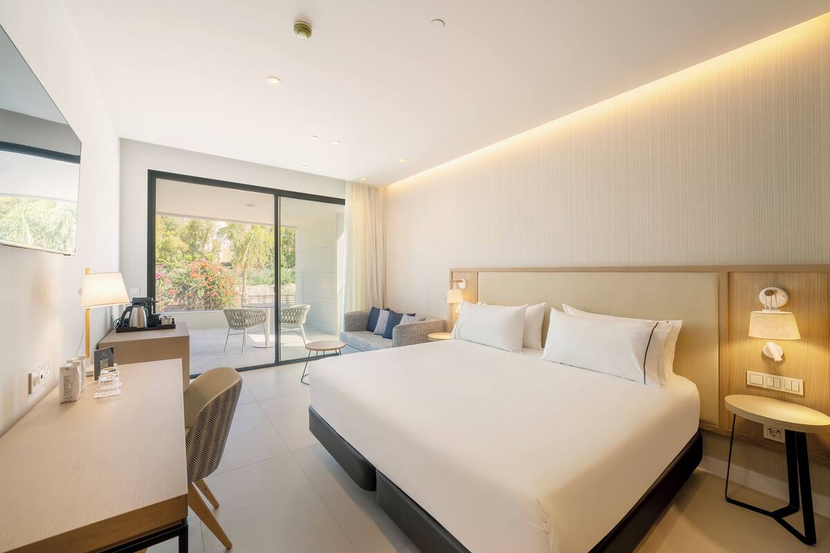 A modern hotel room with a large bed, a desk with a lamp, a chair, and a small table.