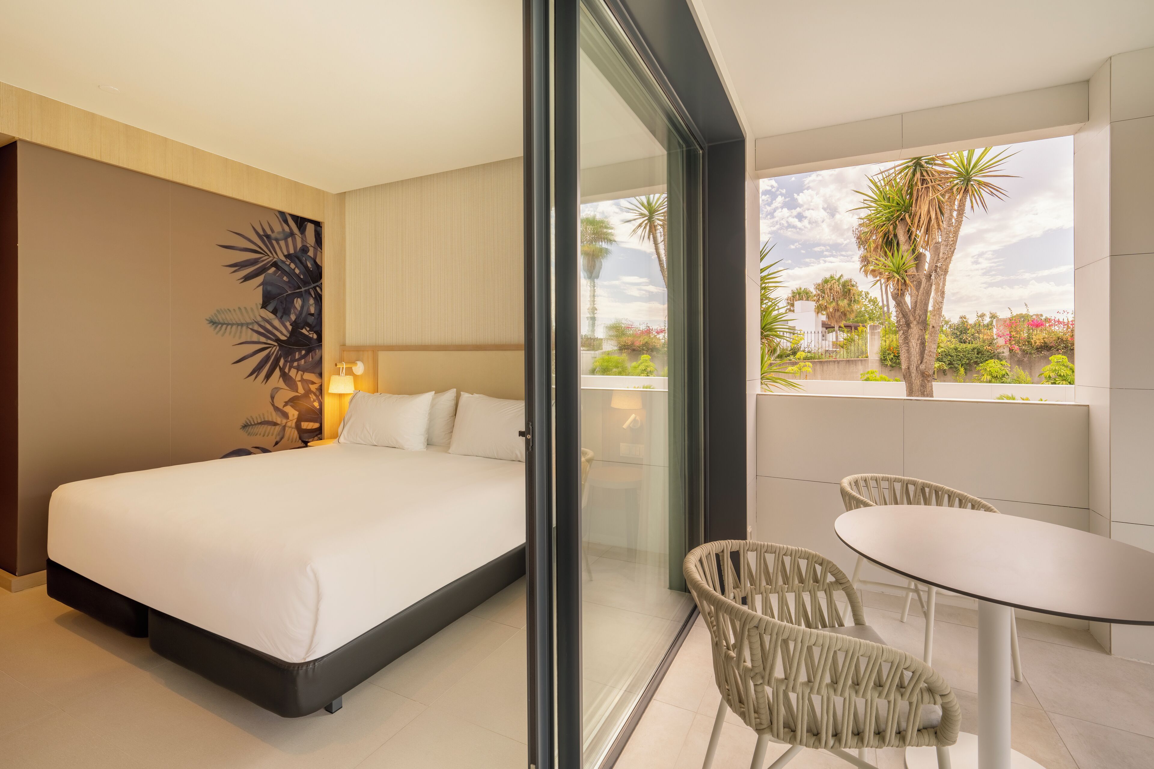Una habitación de hotel moderna con una cama grande, un ventanal de vidrio que da a un balcón con mobiliario exterior y vistas a palmeras.