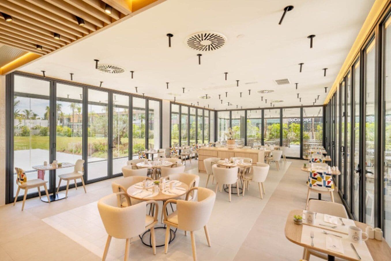 A modern dining area with round tables and chairs, large windows, and a ceiling with recessed lighting.