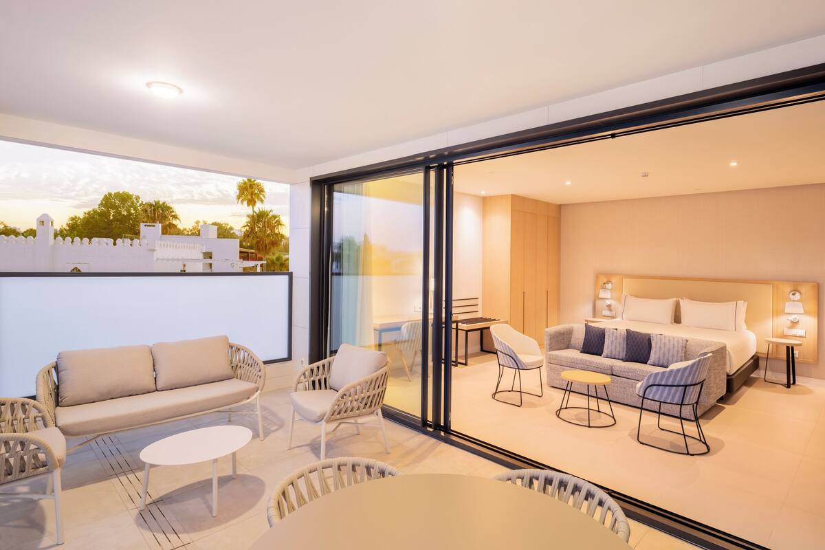 A modern hotel room with a balcony, a sofa, two chairs, a small table, and a bed with a nightstand.