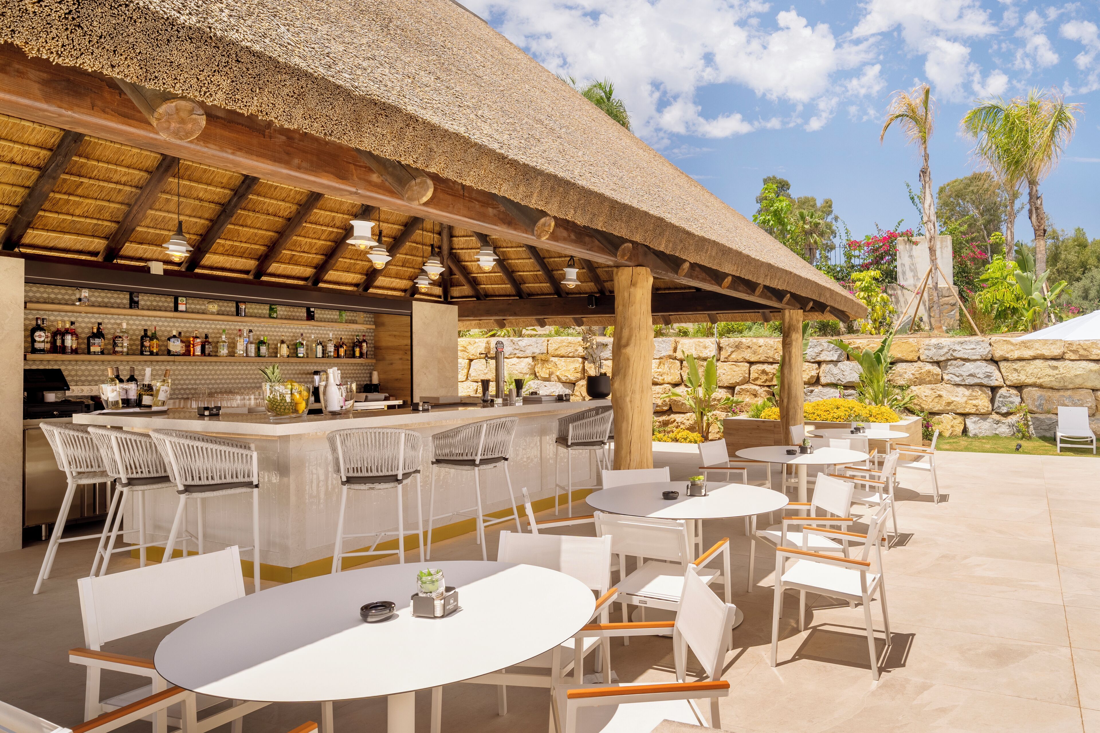An outdoor bar area with a thatched roof, white chairs, and tables. There are bottles and glasses on shelves, and a stone wall in the background.