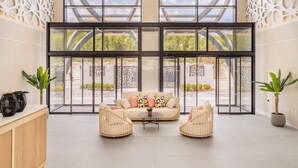 Un vestíbulo amplio con un gran ventanal que da al exterior, un sofá color beige con cojines de colores, dos sillones de mimbre y plantas en macetas.