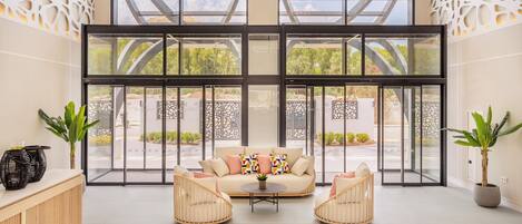 Un vestíbulo amplio con un gran ventanal que da al exterior, un sofá color beige con cojines de colores, dos sillones de mimbre y plantas en macetas.