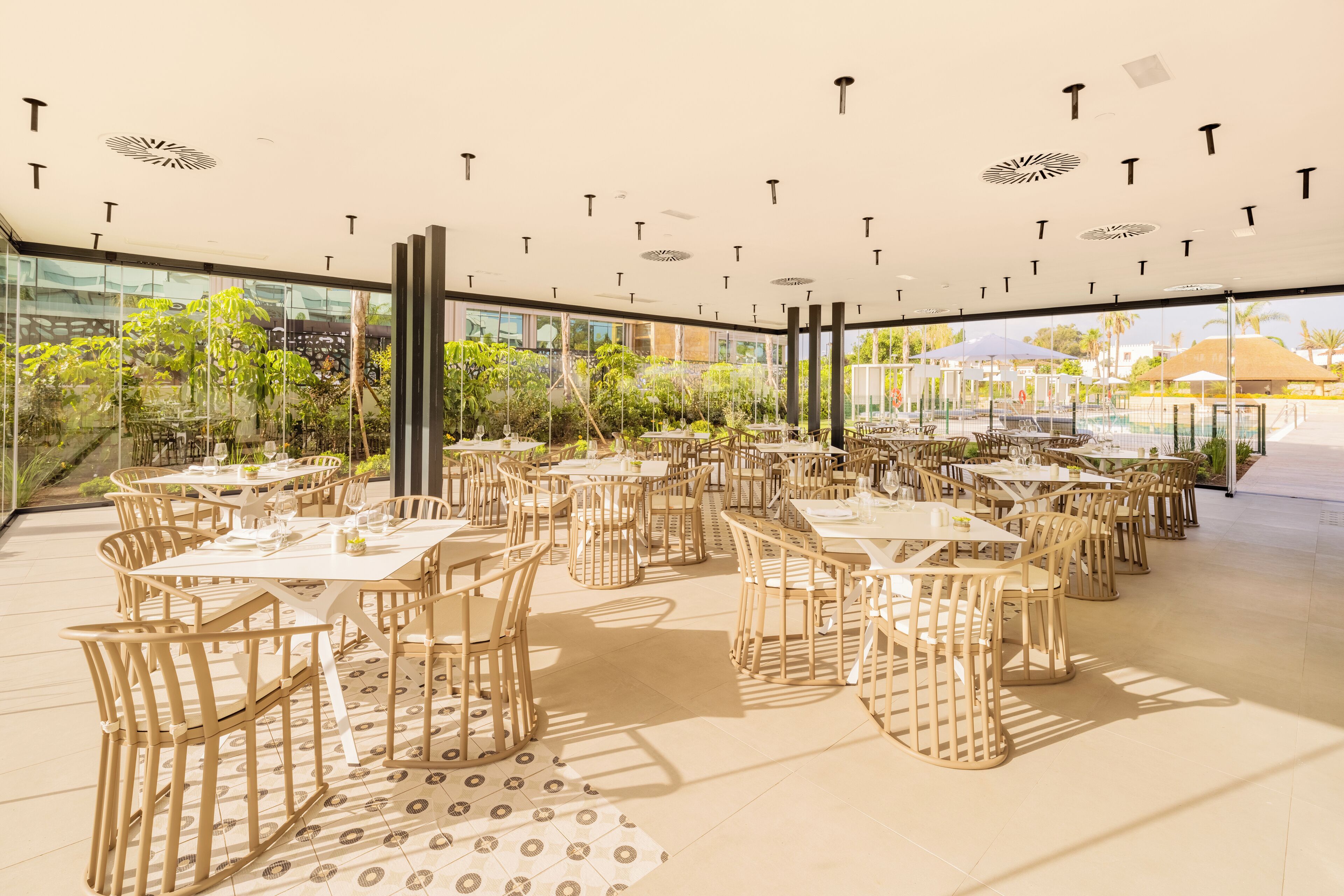 Un área de comedor al aire libre con varias mesas y sillas, rodeada de paredes de vidrio y vistas a una piscina y follaje.