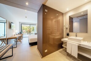 A modern hotel room with a large bed, a desk, a glass-enclosed shower, and a bathroom with a large mirror and sink.