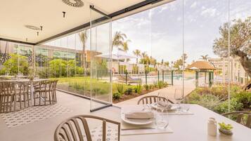 A dining area with a glass wall overlooking a pool and garden. Tables are set with white tablecloths and chairs.