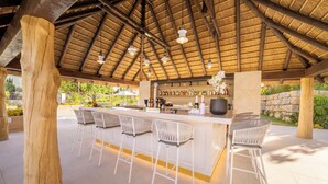 Un área de bar al aire libre con techo de paja, taburetes blancos y una variedad de botellas y vasos en la barra.