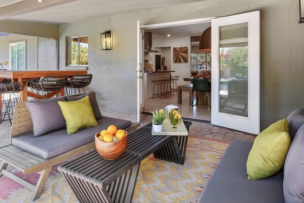 A modern living area with a sofa set, a coffee table with a bowl of oranges, and a dining area visible through an open door.