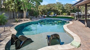 A swimming pool with a curved edge, surrounded by a brick patio. There are two lounge chairs and a covered seating area with tables and chairs. - Glamorous Renovated Desert Oasis, Heated Pool, Tub (Scottsdale)