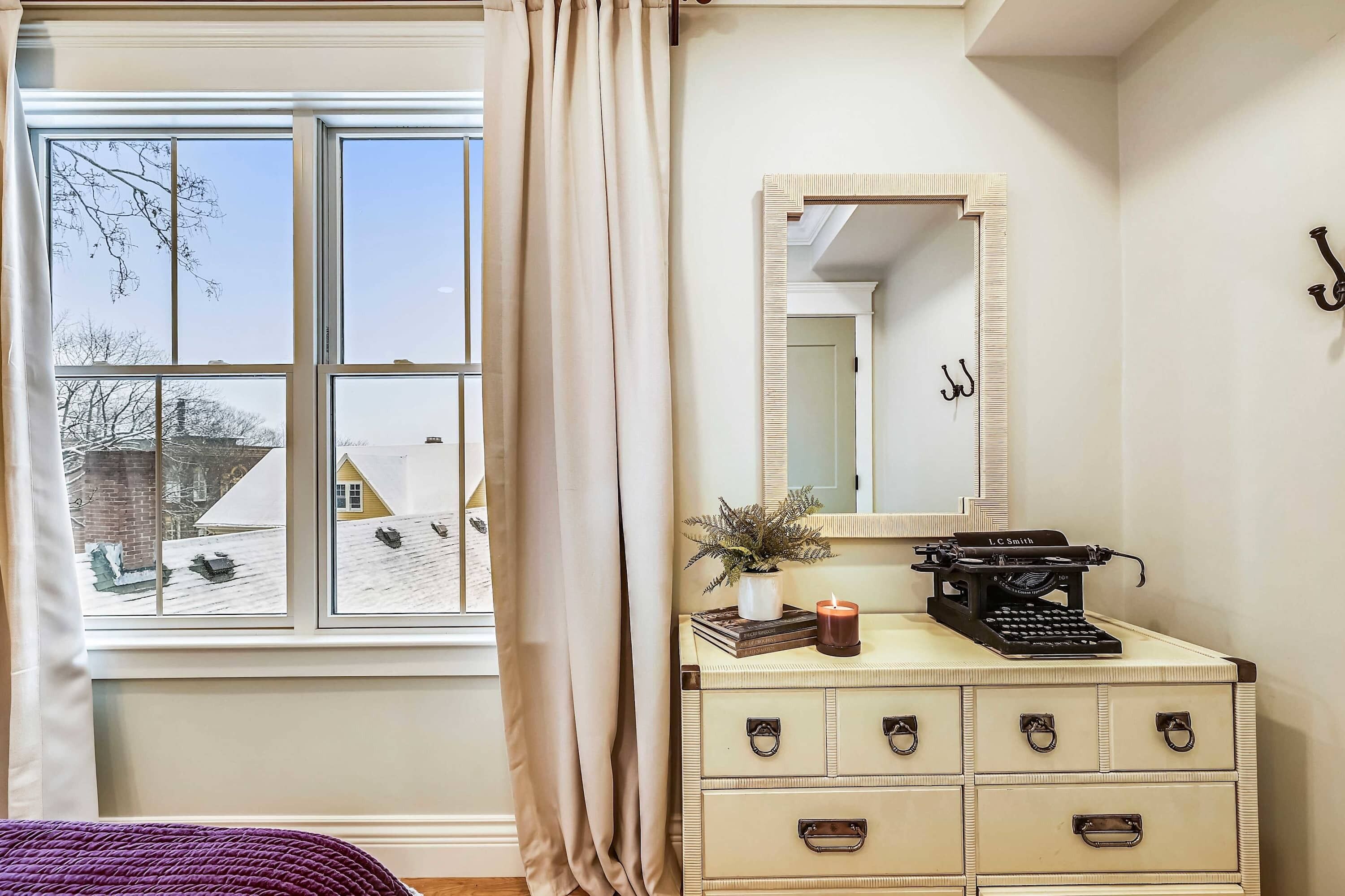 A room with a large window, a dresser with drawers, a mirror, and a typewriter.