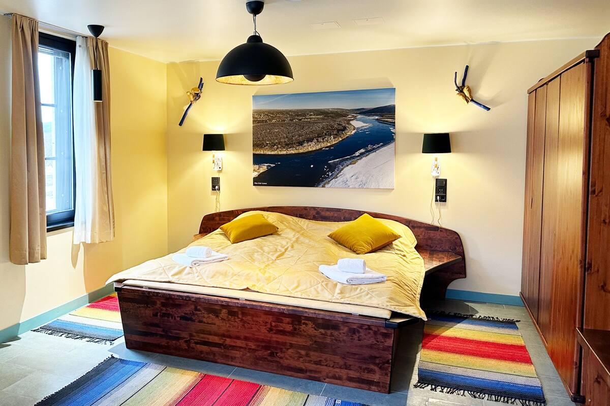 A bedroom with a wooden bed, two bedside lamps, a large landscape painting, and a colorful striped rug.