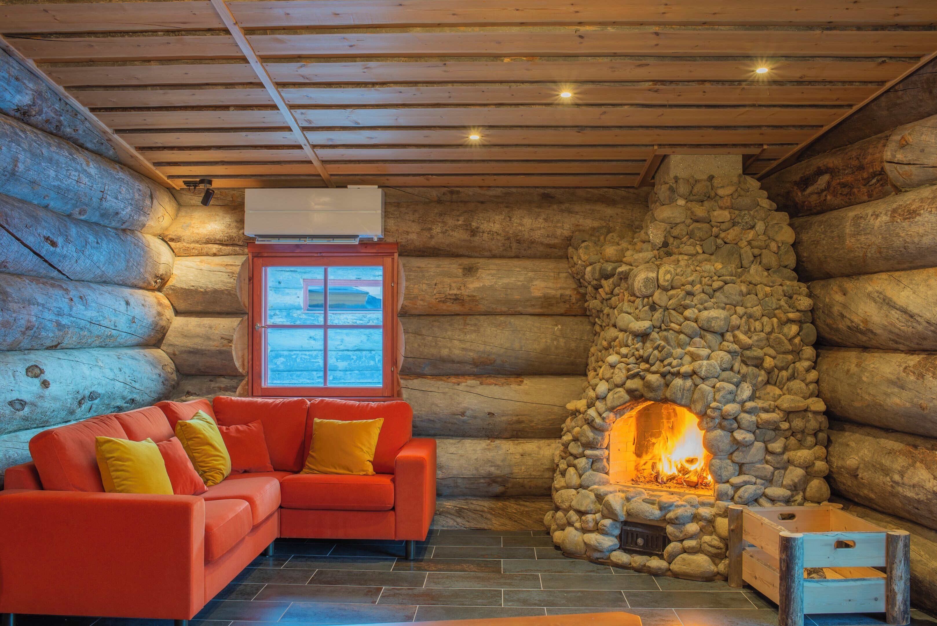 A log cabin interior with a stone fireplace, a red sofa with yellow cushions, and a window with a view.