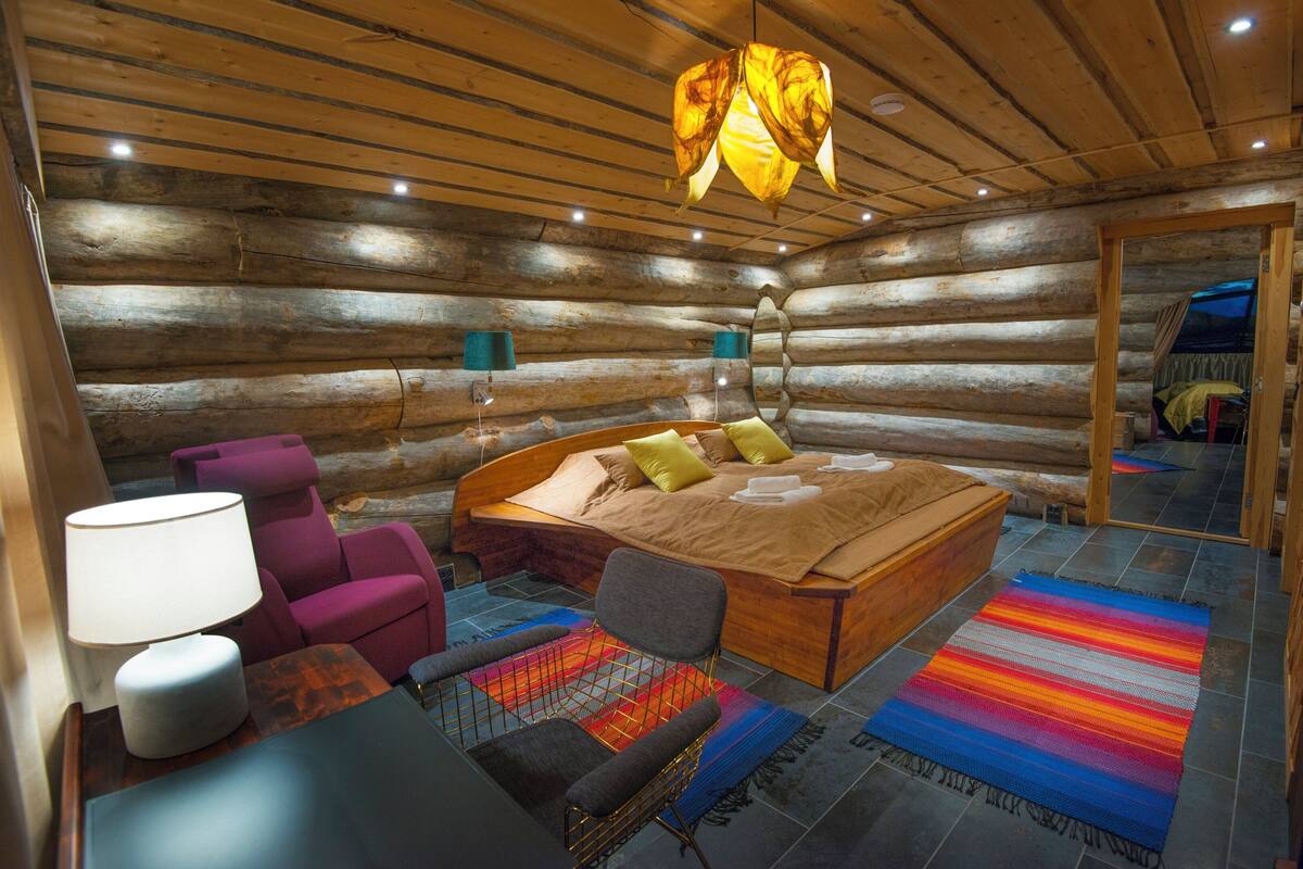 A log cabin bedroom with a wooden bed, a purple chair, a small table with a lamp, and a colorful rug.
