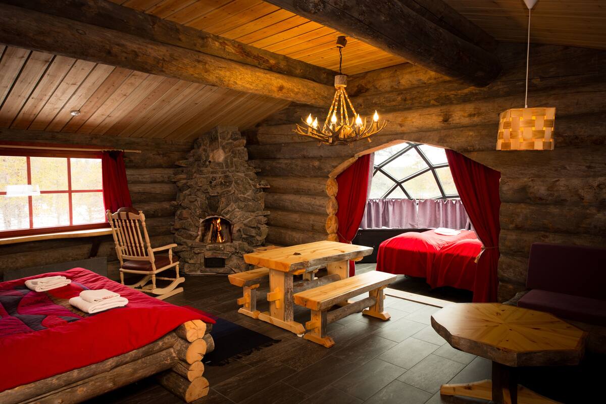 A log cabin room with a stone fireplace, wooden furniture, and a large window with red curtains.