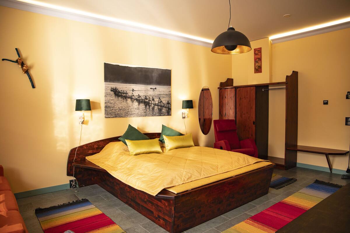 A bedroom with a large bed, a wooden headboard, a colorful rug, a wall-mounted picture, and a hanging light fixture.