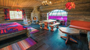 A log cabin interior with a stone fireplace, wooden furniture, and a large window with a view. - Utsjoki Arctic Resort (Utsjoki)