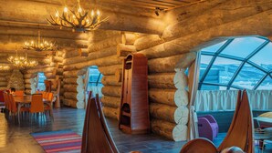 A log cabin interior with a dining area, wooden chairs, chandeliers, and a large window offering a view. - Utsjoki Arctic Resort (Utsjoki)