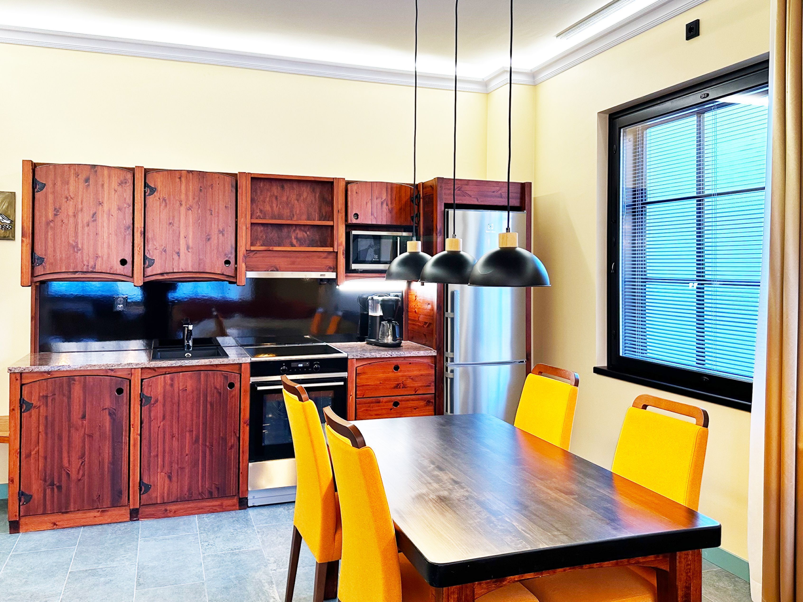 A modern kitchen with wooden cabinets, a stainless steel refrigerator, a built-in oven, and a dining table with yellow chairs.
