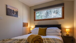 En snyggt bäddad säng med en rutig pläd och en hoprullad handduk, två sänglampor, ett fönster med utsikt över träd och en tavla på väggen.