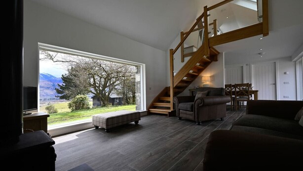 A spacious living room with a large window, a wooden staircase, and a cozy seating area. - The Barn in Banavie near Fort William (Fort William)