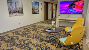 Un hall de hotel con una alfombra estampada, un televisor mostrando una escena colorida y una zona de estar con sillas amarillas.
