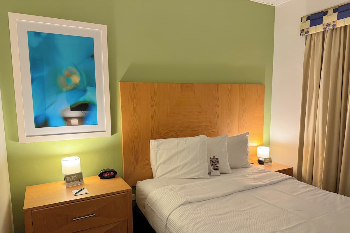 Ein Hotelzimmer mit hölzernem Kopfteil, einem Bett mit weißer Bettwäsche, einem Nachttisch mit Lampe, einem gerahmten Bild an der Wand und einem Beistelltisch mit Uhr und Schilder.