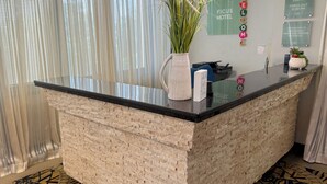 Un mostrador de recepción de hotel con un muro de acento de ladrillo, un jarrón con plantas y un cartel de registro de entrada y salida.