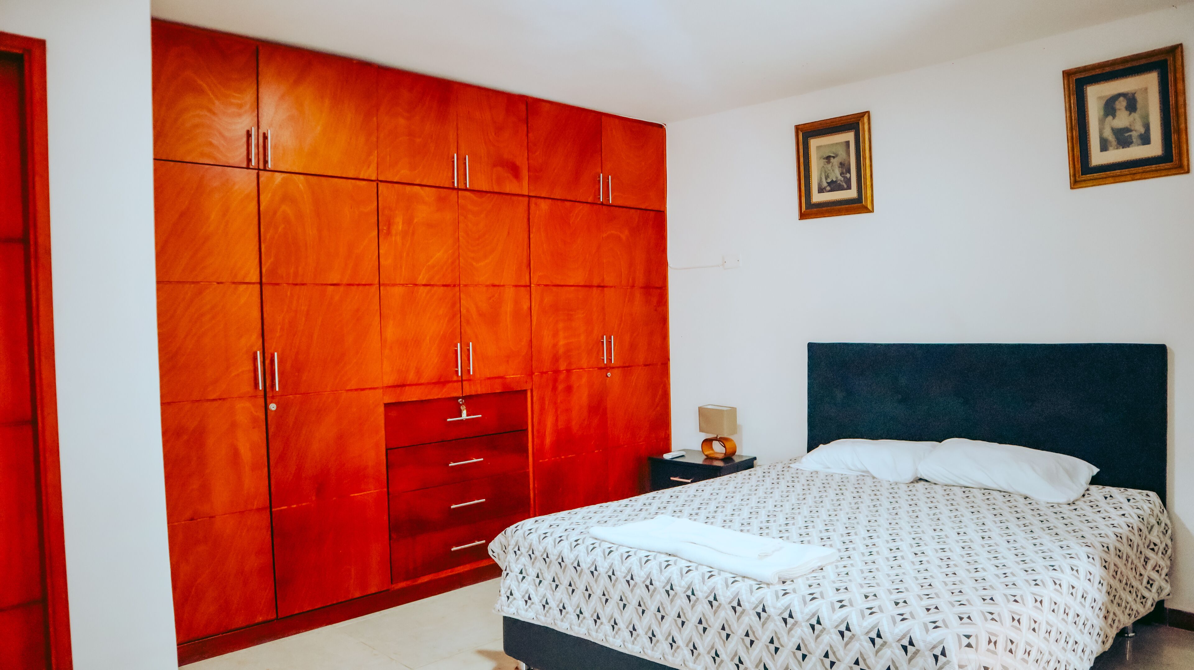 Un dormitorio con un amplio armario de madera, una cama con colcha estampada y dos cuadros en la pared.