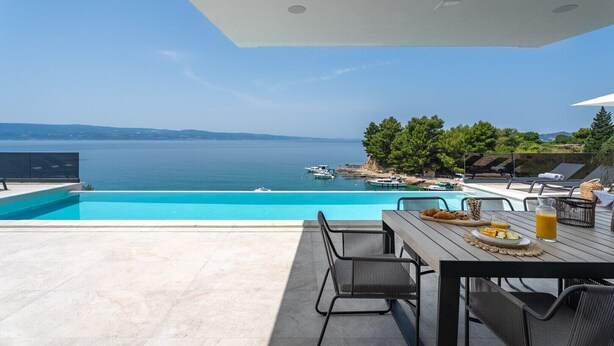 Un espace aménagé au bord de la piscine, avec vue sur la mer, des sièges d’extérieur et une table dressée avec de la nourriture et des boissons.