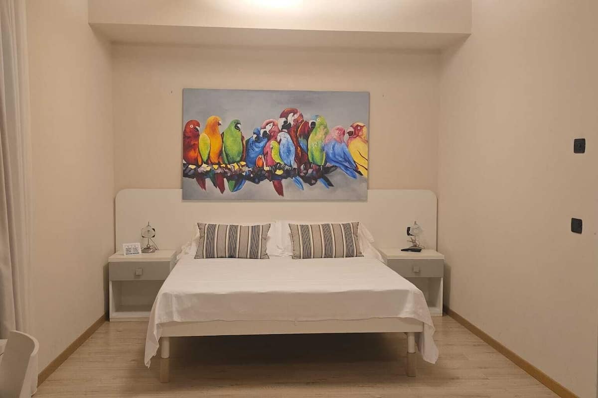 A bedroom with a bed, two bedside tables, a nightstand, a painting of colorful birds, and a wooden floor.