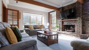 A living room with a brick fireplace, a television, and a large window. - Gracelands: Swiss Inspired Home (Blackstone Heights)