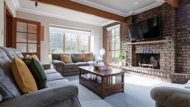 A living room with a brick fireplace, a television, and a large window. - Gracelands: Swiss Inspired Home (Blackstone Heights)