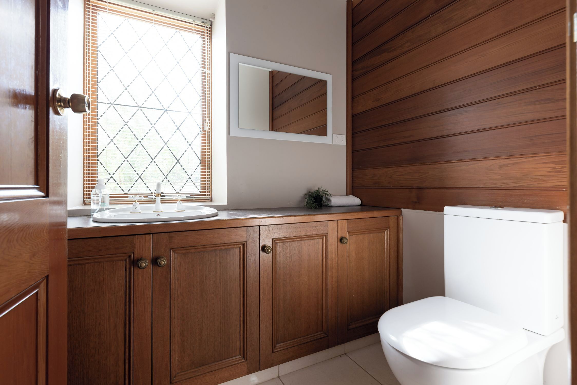 A bathroom with wooden cabinets, a toilet, a sink with a mirror, and a window with blinds.