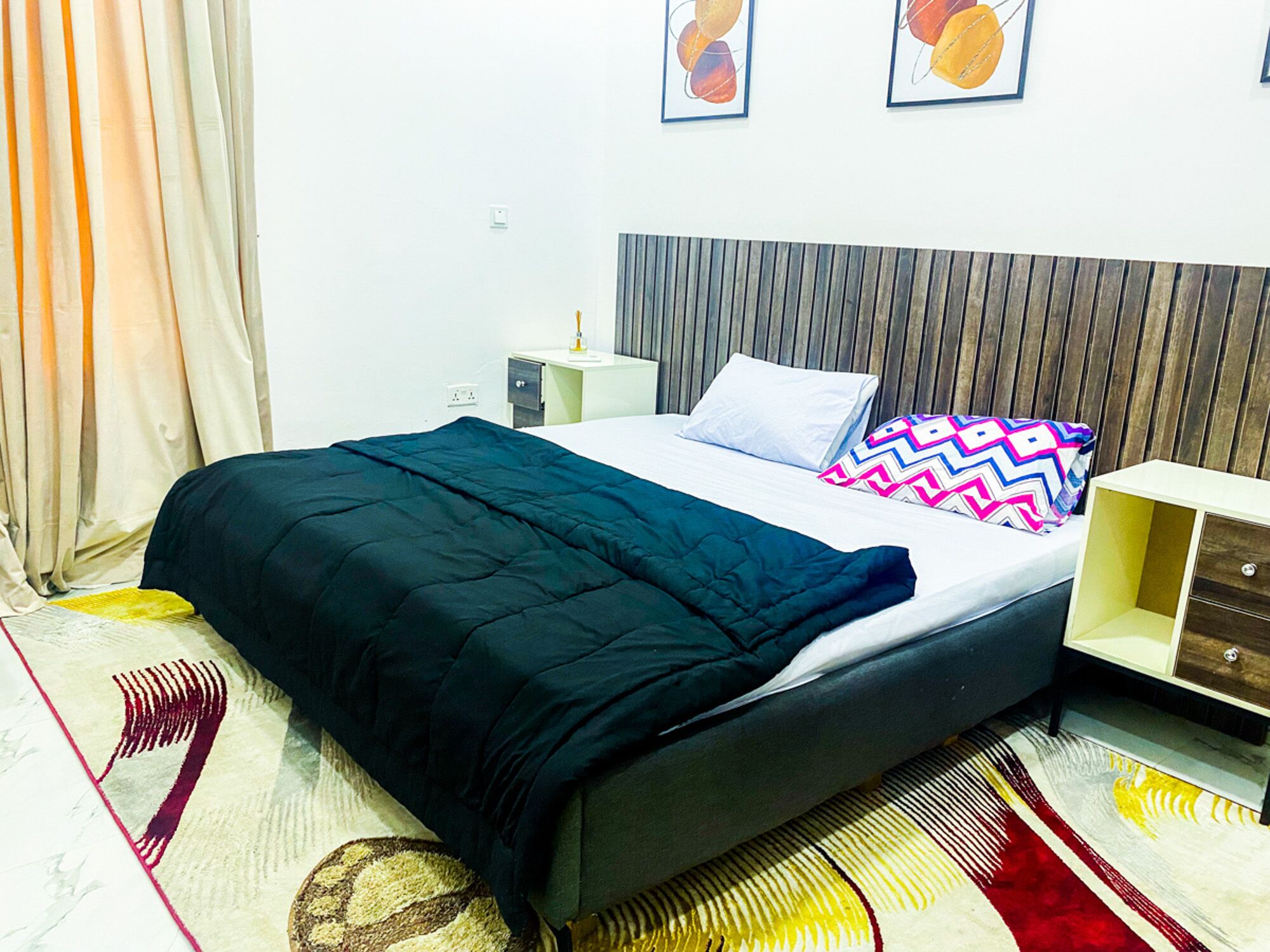 Una cama bien hecha con edredón negro, un cabecero de madera y un cojín colorido. Hay una mesita de noche con una lámpara y un cajón pequeño. La habitación tiene una pared blanca y una alfombra estampada.