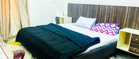 Una cama bien hecha con edredón negro, un cabecero de madera y un cojín colorido. Hay una mesita de noche con una lámpara y un cajón pequeño. La habitación tiene una pared blanca y una alfombra estampada.