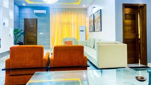 A modern living room with a glass coffee table, orange armchairs, a beige sofa, and a chandelier. - Impeccable 2-bed Apartment in Lekki With Snooker (Lekki)