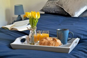 Un plateau de petit-déjeuner sur un lit, avec un livre, un vase de fleurs jaunes, un verre de jus d’orange, une tasse et un croissant.