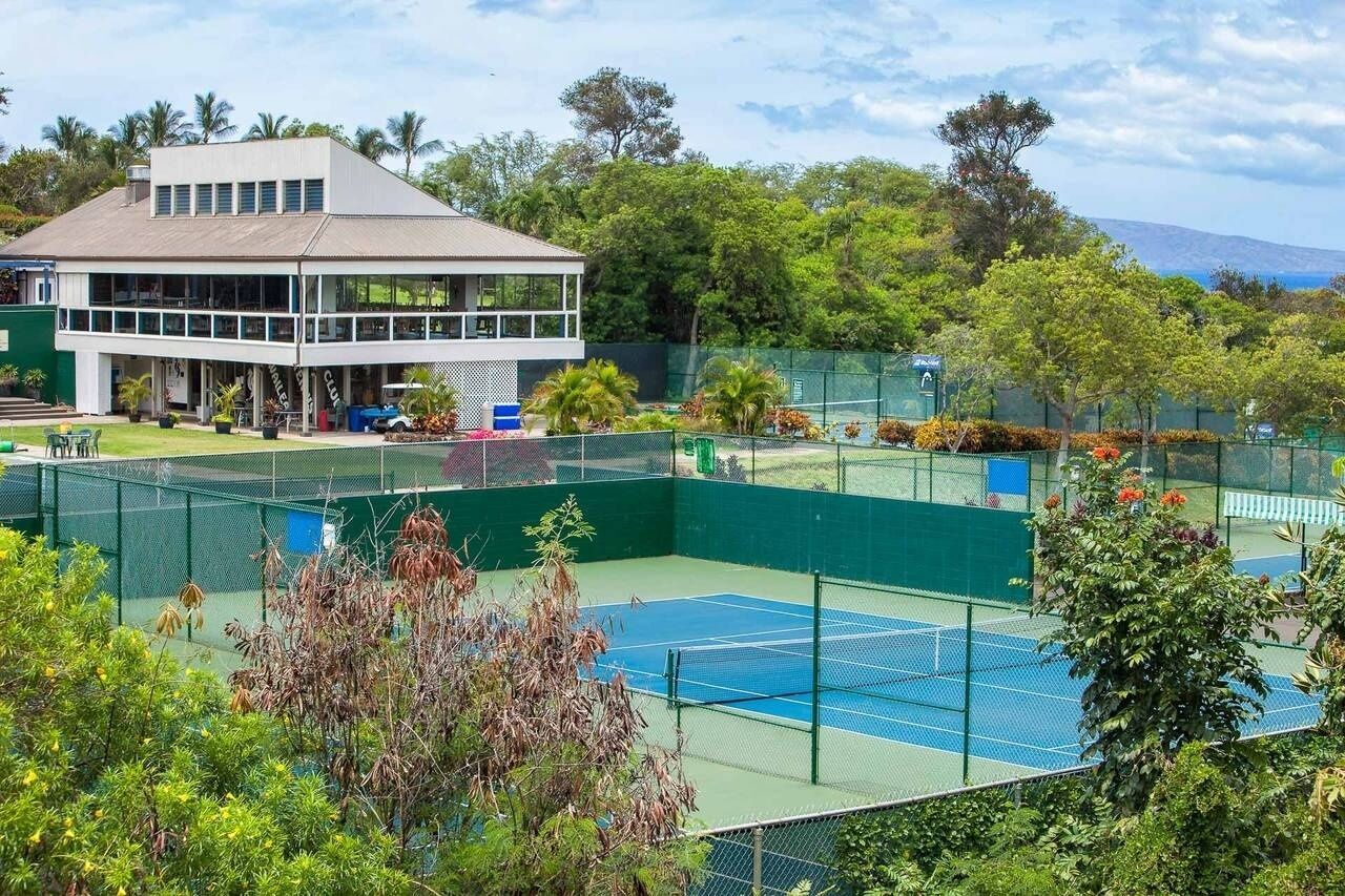 Un court de tennis entouré de verdure, un bâtiment doté d’un toit plat et un paysage montagneux en arrière-plan.