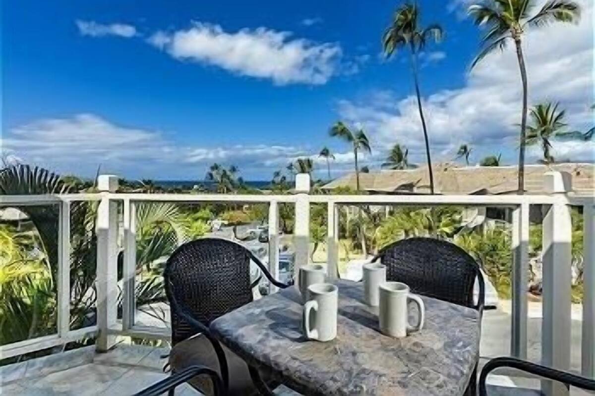 Eine Terrasse mit zwei Stühlen und einem Tisch, mit Blick auf einen Strand mit Palmen und einem klaren Himmel.