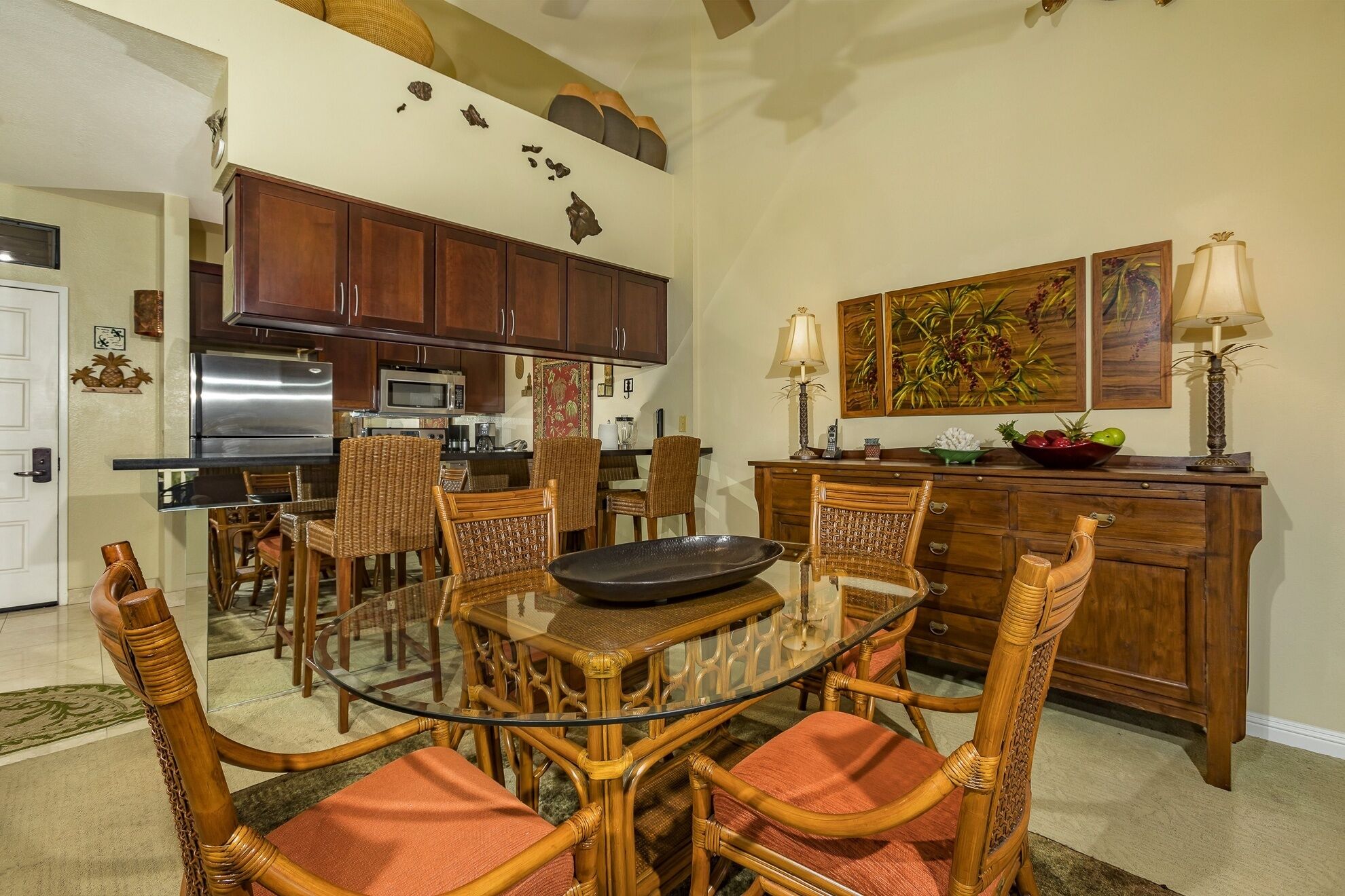 A dining area with a glass table surrounded by wicker chairs, a kitchen in the background, and artwork on the walls.
