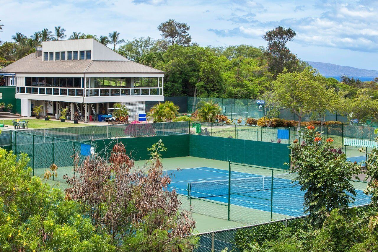 Un court de tennis avec un filet, entouré d’une clôture verte et d’arbres.