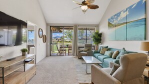 A spacious living area with a large TV, comfortable seating, a ceiling fan, and a view of the outdoors. - Grand Champions Two Bedrooms - Ocean View by Coldwell Banker Island Vacations (Kihei)