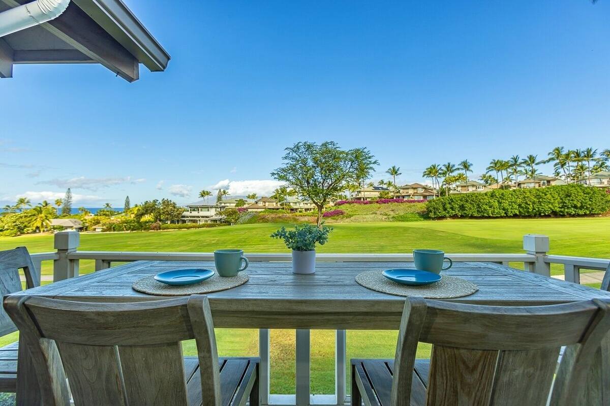 Ein Holztisch mit zwei blauen Tellern und zwei blauen Tassen auf einem Balkon mit Blick auf einen Golfplatz, bewölkter, klarer, blauer Himmel.